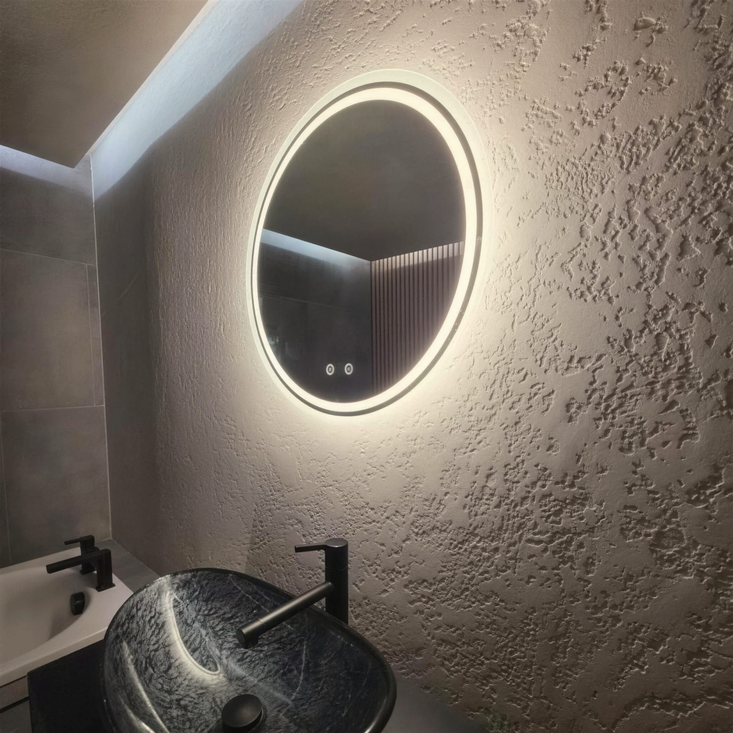 Halo-lit round mirror on a deeply textured plaster wall above a black marbled vessel basin, in cool moody light.