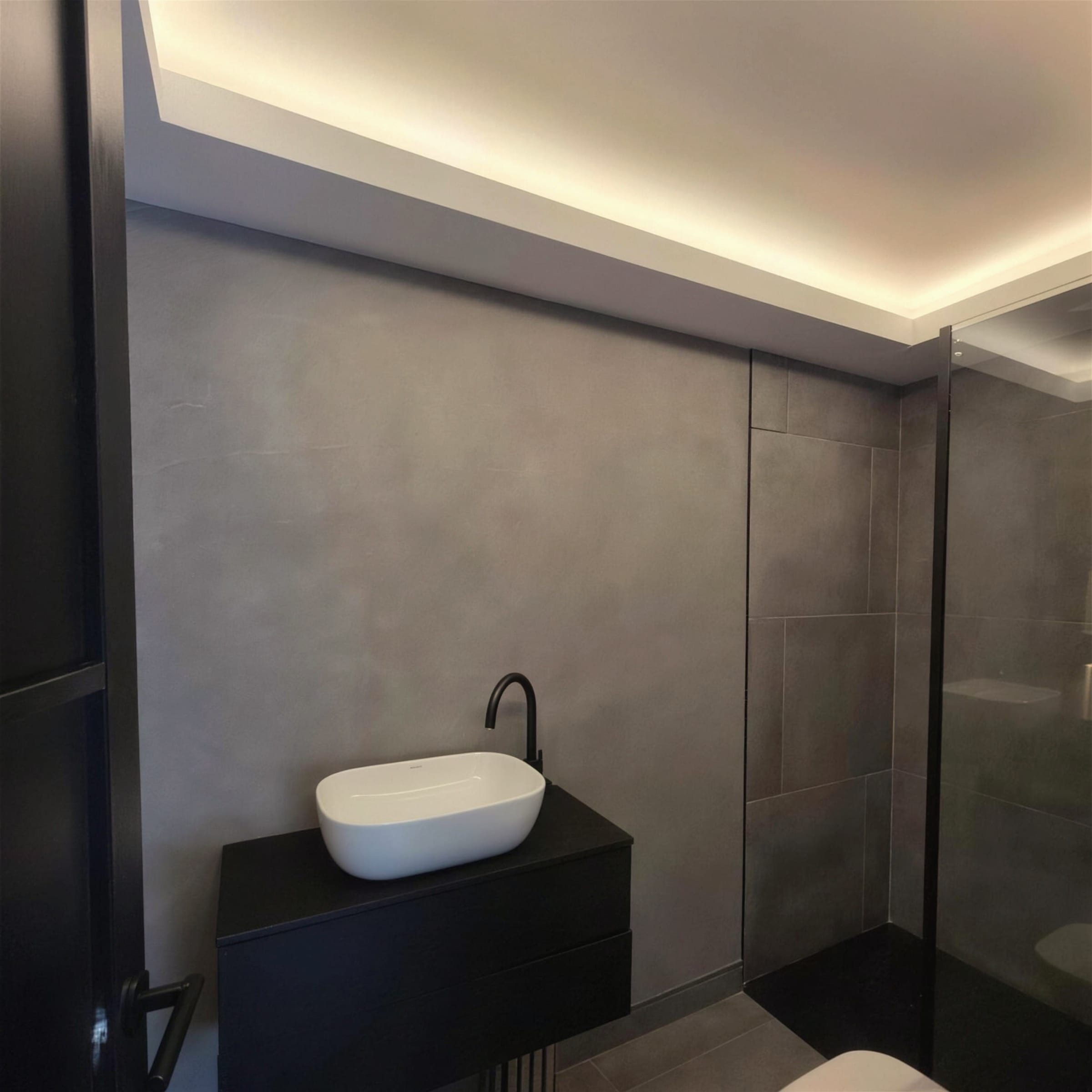 Bathroom vanity wall in grey concrete-effect plaster under a warm LED cove, with a black wall-hung unit and white countertop basin.