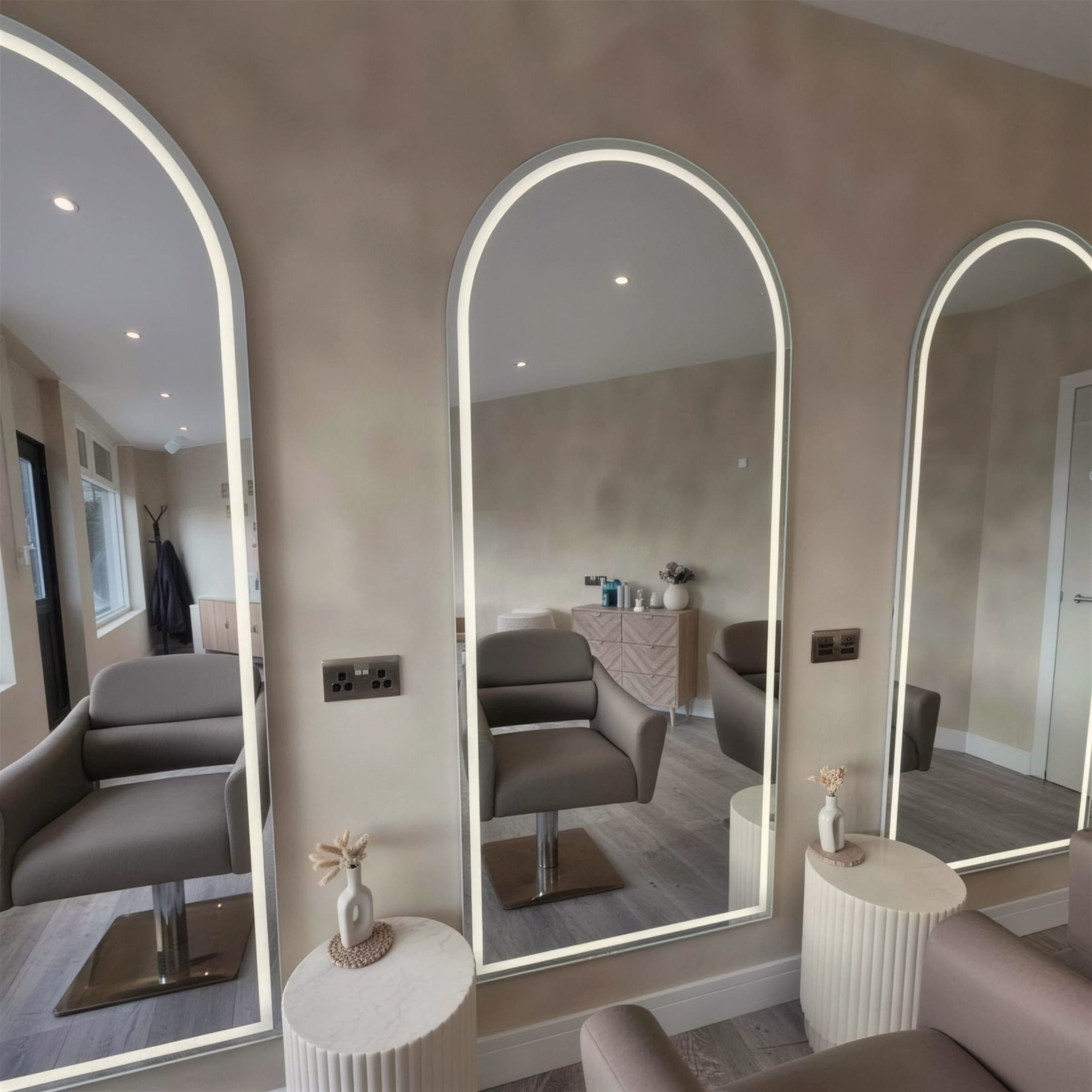 Bright salon interior where limewash plaster walls set off a row of arched LED mirrors, styled stations and taupe chairs.