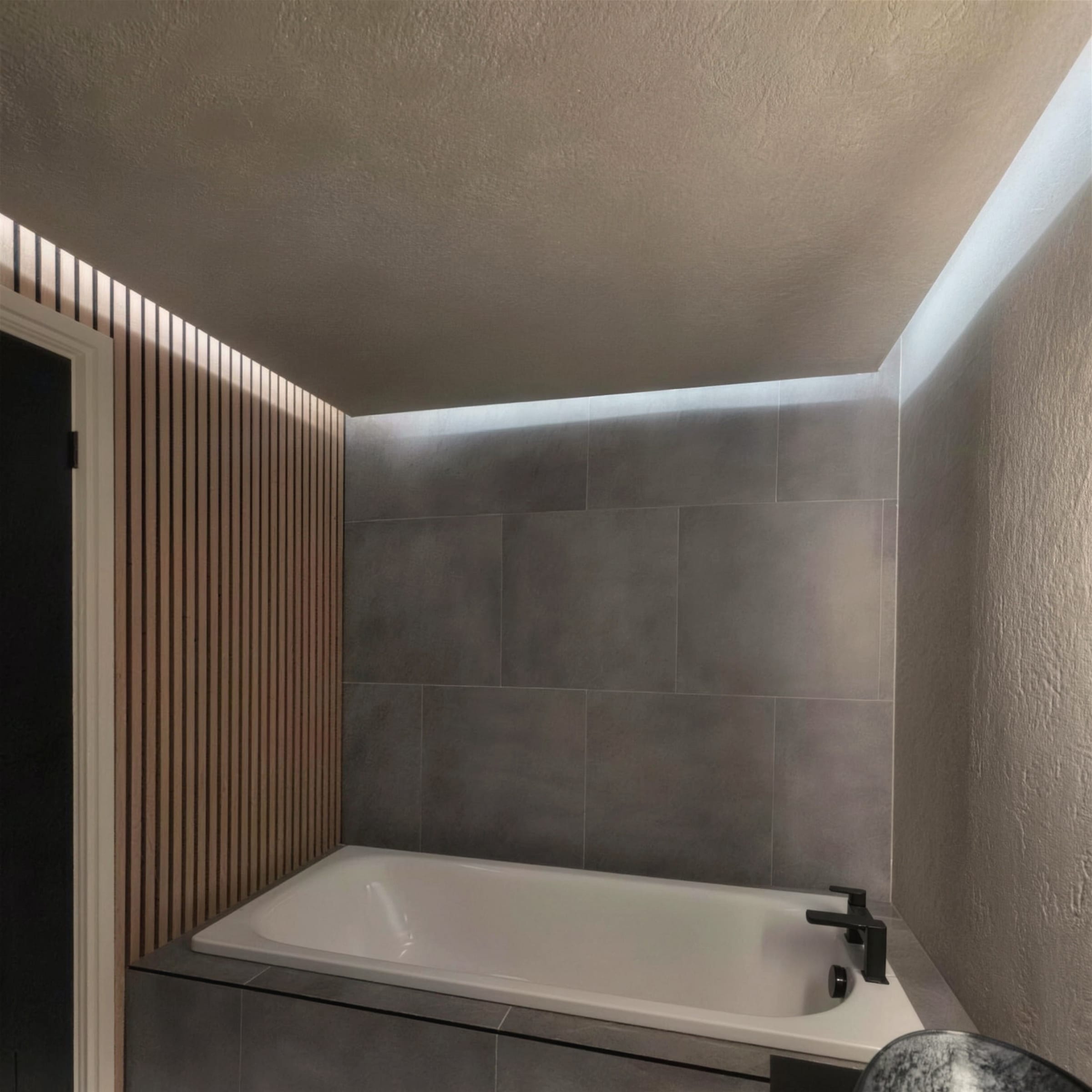 Moody bath corner combining grey tiles, warm wood slats, matt-black tap and a rustic textured plaster ceiling washed by an LED cove.