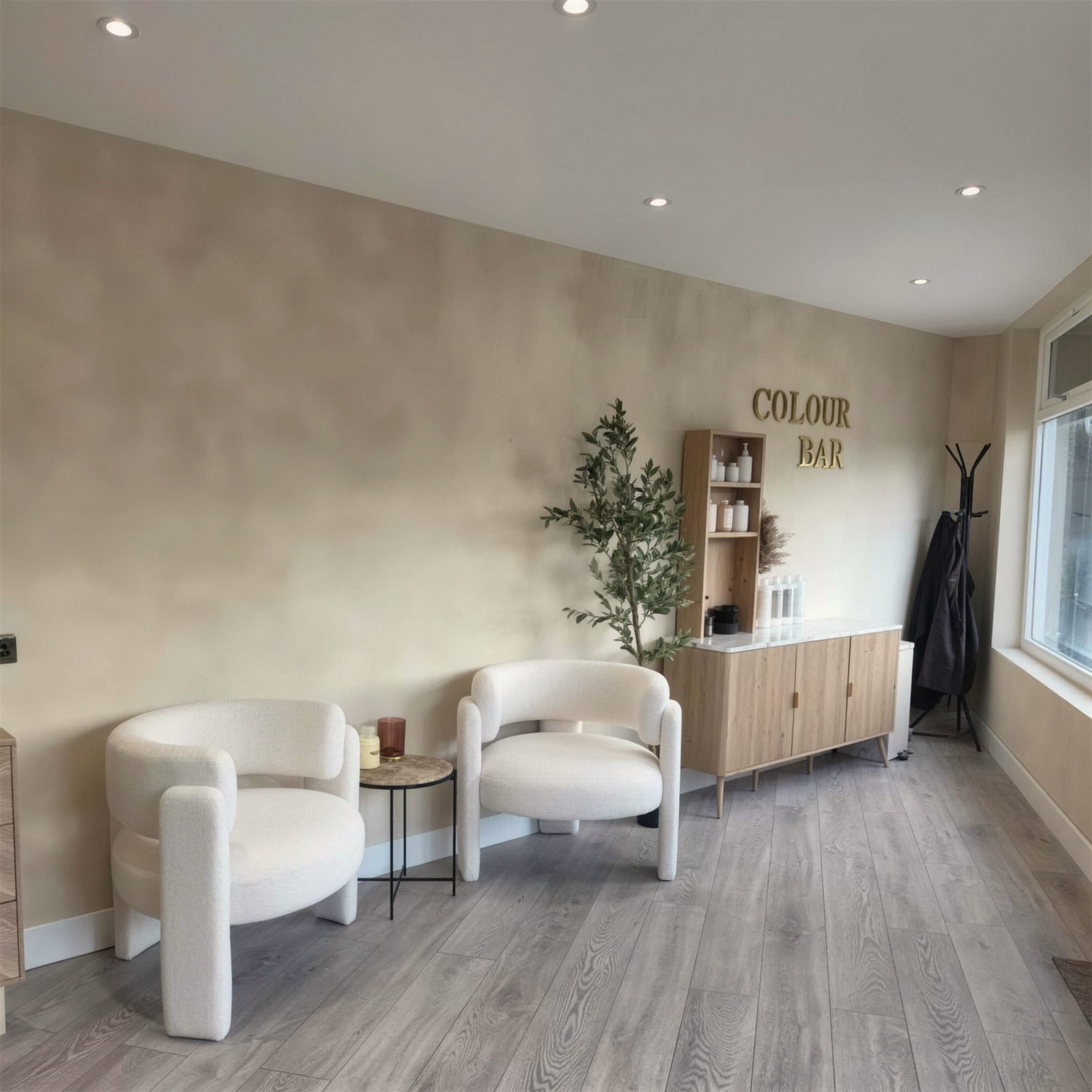 Salon waiting corner with two white boucle chairs, olive tree and oak cabinet under gold Colour Bar lettering on limewash plaster.