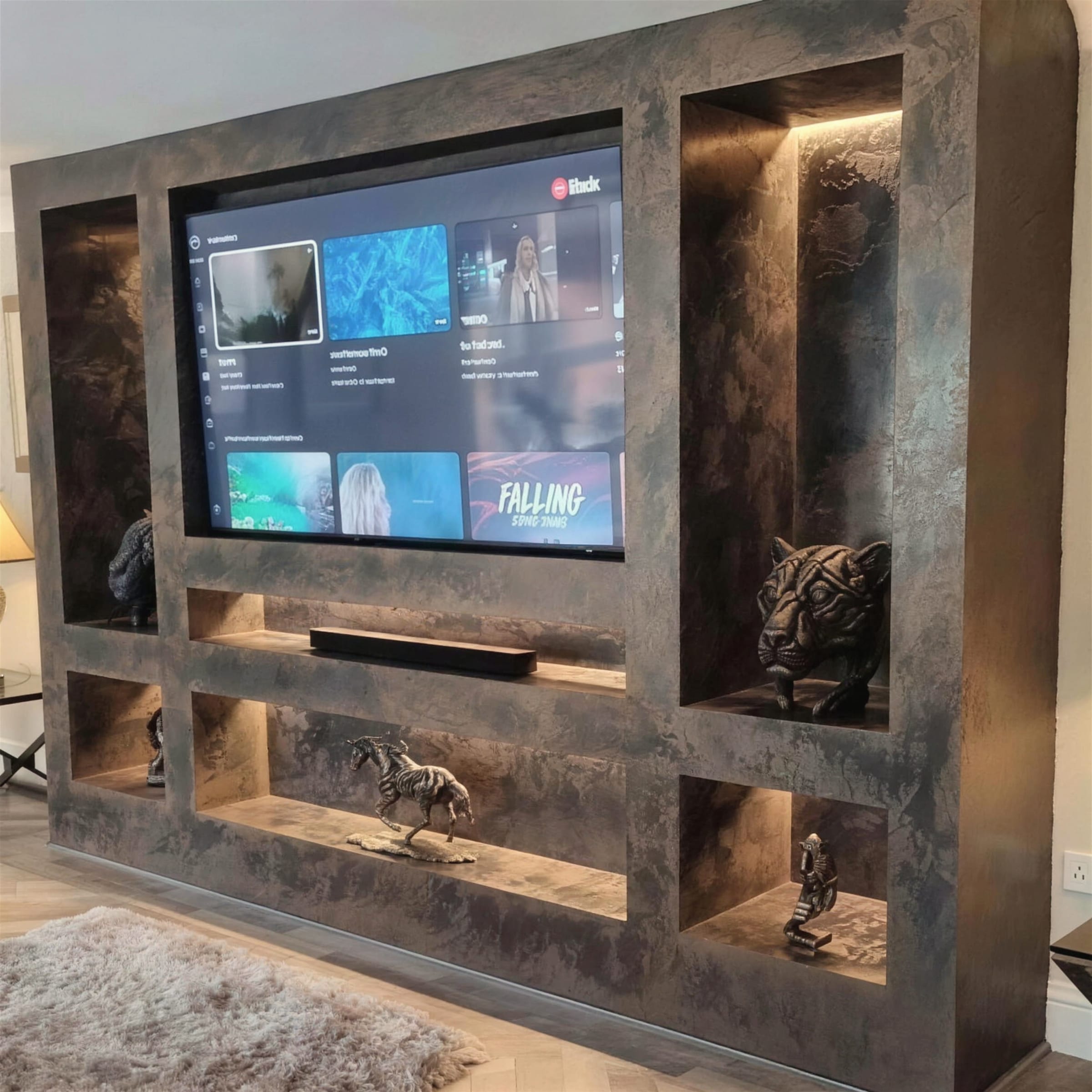 Bespoke media wall finished in dark oxidised metallic plaster with warm-lit display niches holding bronze animal sculptures around a wall-mounted TV.