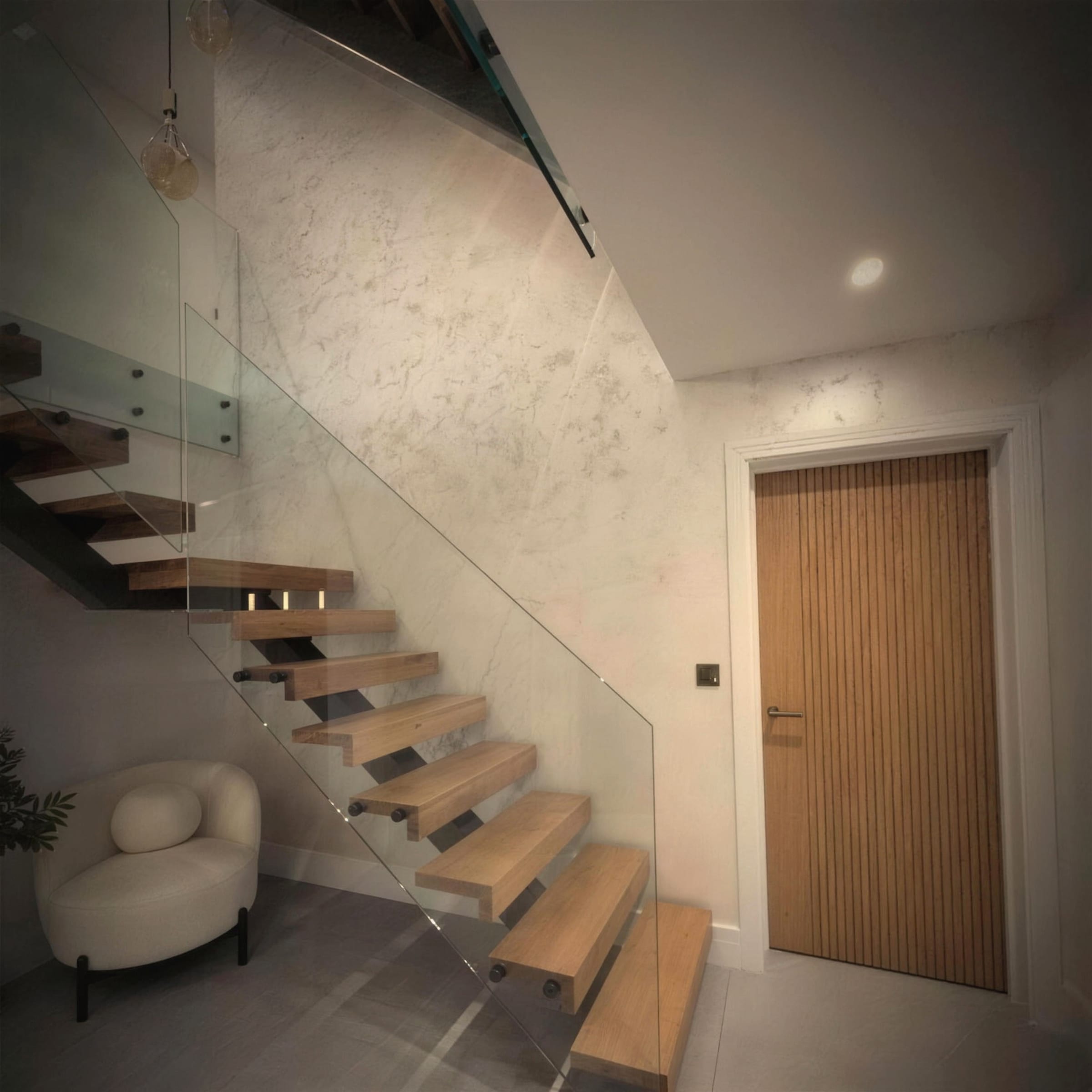 A floating oak staircase with a frameless glass balustrade rises along a softly mottled cream polished plaster wall, warmly lit, with an oak slatted door and a boucle armchair beneath.