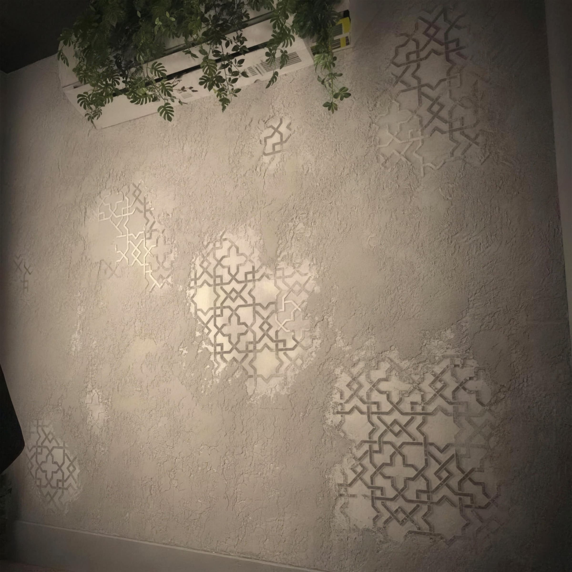 A rustic textured plaster feature wall in warm greige with geometric Moorish stencil motifs emerging in smooth relief, softly raked light, trailing greenery at the top.