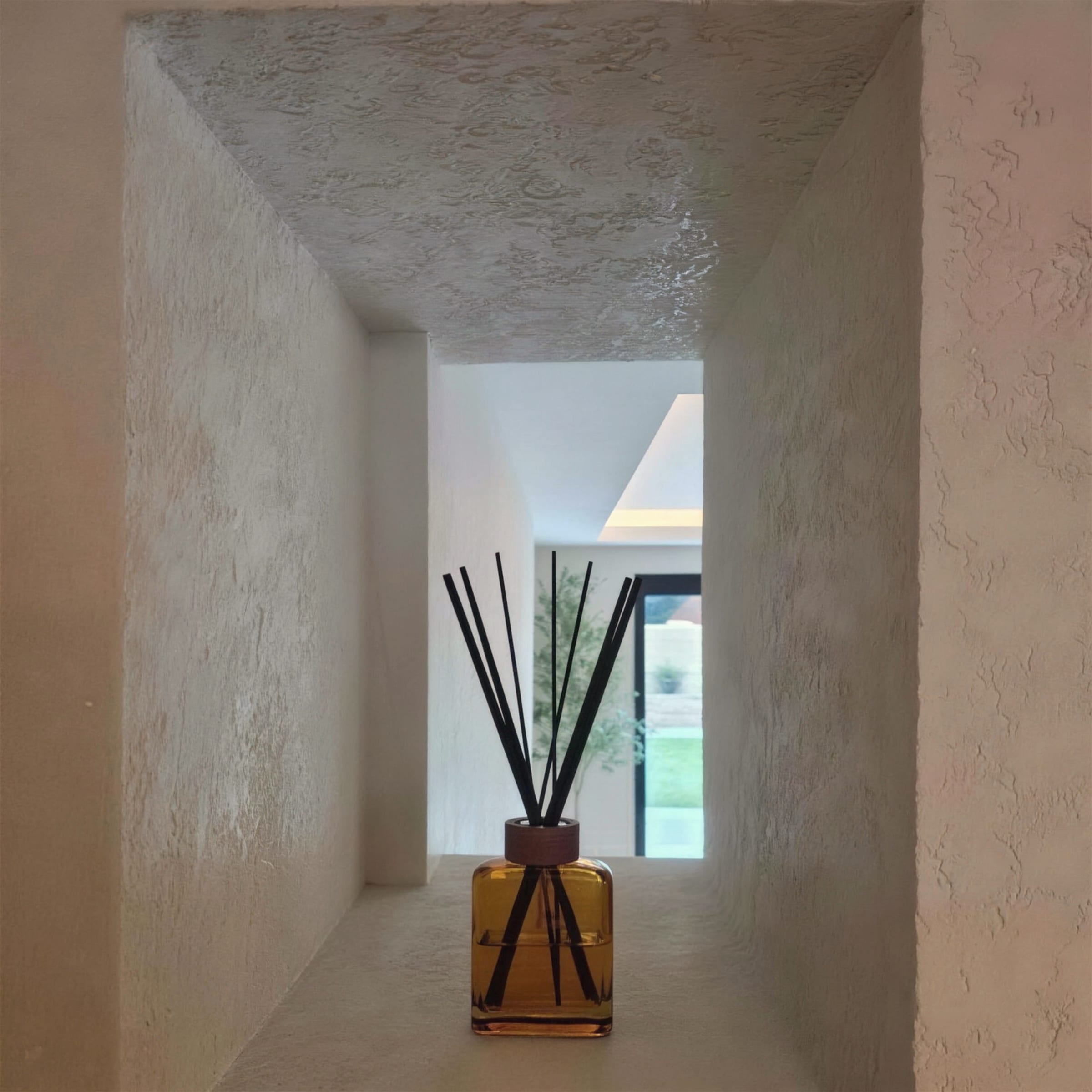 Deep cream textured plaster niche framing an amber reed diffuser, layered view through to a bright modern interior and garden beyond.