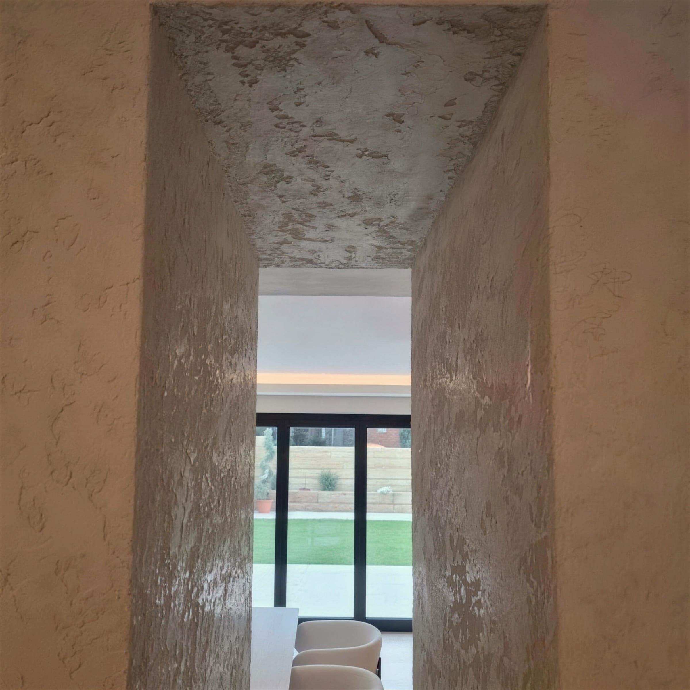 Doorway passage fully clad in heavily textured rustic plaster, walls and ceiling reveal included, looking through to glazed doors and a green garden.