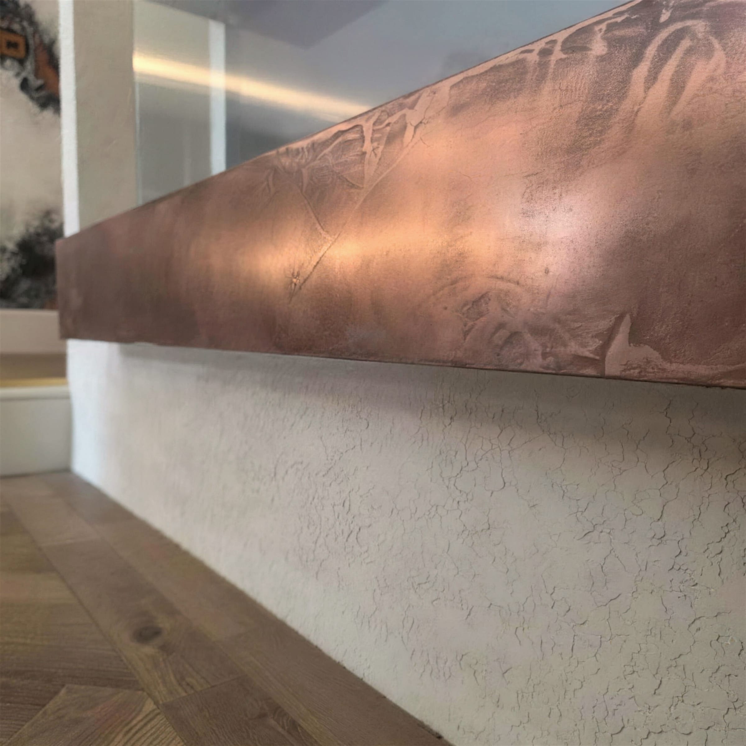 Close diagonal detail of a burnished liquid-copper metallic plaster beam above a white textured plinth and herringbone oak floor.