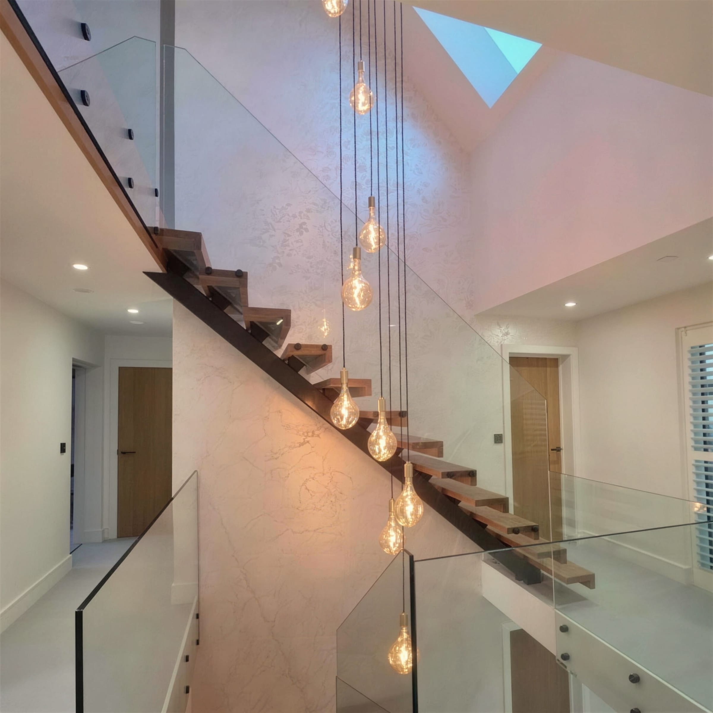 Double-height stair atrium with floating oak treads, glass balustrade and a cascade of filament pendants against seamless cream polished marmorino walls under a skylight.